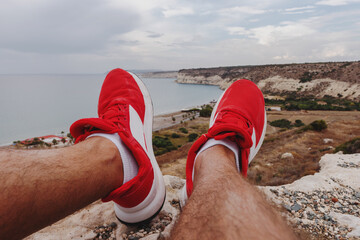 First-person perspective of person is wearing red sneakers resting on a rocky cliff with a coastline view.