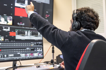 A television director in a broadcast control room, actively guiding a live production. Surrounded by multiple screens, the director wears a headset and gestures towards a video switcher.