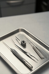 A close-up of surgical tools arranged on a metal tray, including a scalpel, tweezers, and forceps, set in a clinical environment.