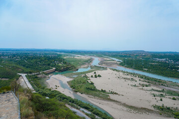 A high-angle view of a winding river flowing through a vast landscape with green vegetation, sandbanks, and distant settlements under a clear sky.