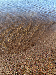 Seafoam covers the colorful small shells on the beach. A sunny day by the sea. Little sea shells, glass, and smooth stones lay on the water's sand. Closeup photo. Walking and noticing. 