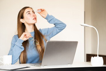 A young woman using medicine eye drops while sitting at desk taking a break from work on laptop....