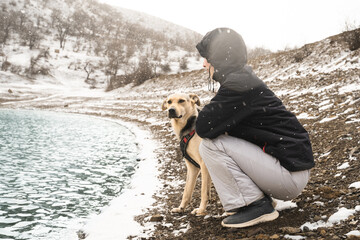 Dog and its woman owner sitting by a turquoise mountain lake on a snowy sunny winter day. Human-animal bond, companionship, adventure with pet, friendship, exploration, hiking, walk, journey.