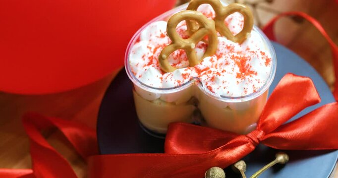 Heart shaped dessert cup layering cake and cream, topped with whipped cream, crunchy pretzels, red sprinkles, sitting on dark plate beside crimson ribbon