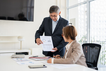 Senior professionals work on project in modern office, analyzing documents and charts. The man focuses intently the woman reviews papers with calculator ,dynamic scene of collaboration and expertise.