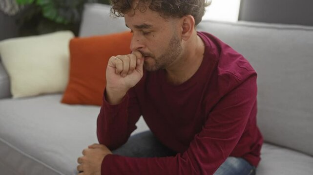 Young man sitting nervously on a couch in a cozy living room with orange and white pillows, reflecting anxiety and contemplation in a homely indoor environment.