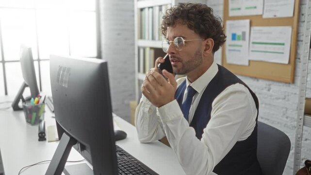 Hispanic man talking on phone in modern office setting with computer and documents emphasizing business communication and professional environment