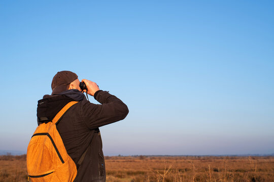 A man in a cap and with a yellow backpack in a field on a sunny day in spring watches birds and wild animals with binoculars against a blue sky. Bird watching