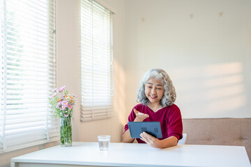Mature 60s woman waving hand hello at tablet screen, speaking on video call, talking to family online, smiling, sitting on sofa at home. Distant communication concept