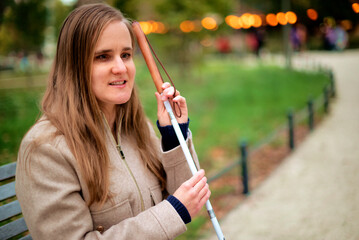 A visually impaired woman with a white cane relaxing on a bench in a park on an autumn day