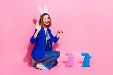 Smiling young woman in a blue jacket posing with playful bunny ears and colorful rabbit figures on a pink background