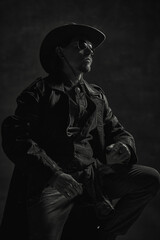 Black and white portrait of strong cowboy in leather coat and hat, hands resting on knees, radiating authority and wisdom under moody lighting. Concept of leadership, western culture, history.