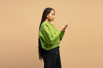 Young woman using mobile phone, standing in profile view on beige background