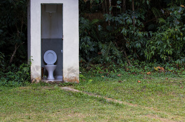country house bathroom in the middle of a grassy area with the forest in the background with an open door and a view of a toilet with an open lid and a wall-mounted flush tank
