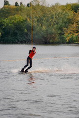 A girl in a red vest rides a wakeboard on a lake against a background of trees, creating splashes...