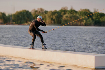 A wakeboarder slides along a figure in a cable park during the day, making hand gestures and...