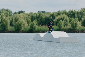 A wakeboarder glides along a white bullock-shaped springboard on a lake against a background of...