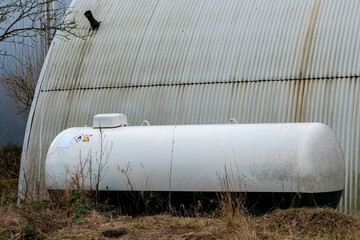 A white storage tank rests next to a corrugated metal building, overgrown with grass and weeds. The...