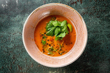 A vibrant bowl of spicy soup garnished with fresh cilantro and lime, set against a textured dark background. The contrasting colors create an appetizing visual.