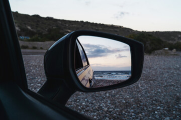 Beach scene is reflected in the side mirror of a parked car, capturing the ocean waves and evening sky