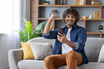 Obraz premium Cheerful man with curly hair celebrating success while using a smartphone at home, sitting comfortably on the sofa.