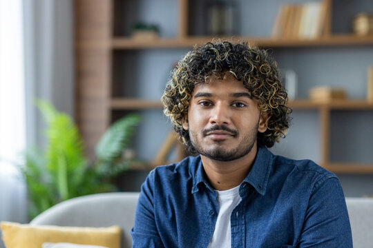 Portrait of a handsome young man with curly hair, wearing a denim shirt, looking directly at the camera in a cozy home setting. - Powered by Adobe