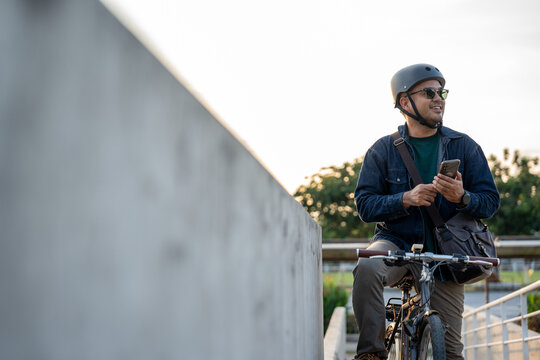 Asian young traveler man riding bike using smartphone navigate the gps map. Sustainable save energy lifestyle. Commute carbon neutral transportation. Tourist with helmet cycling bike to urban city - Powered by Adobe