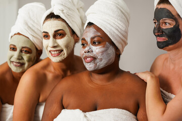 Multiracial women enjoy spa day together with beauty face mask while wearing towels - Multi generational female friends with different skin color and types