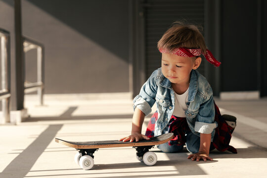 Delightful toddler boy wearing a denim outfit and red bandana plays with a skateboard in an urban setting