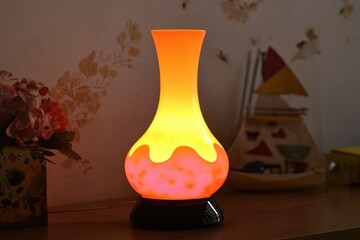 A vibrant lava lamp with a glowing orange and yellow design, creating a warm ambiance, sits on a wooden surface against a subtly decorated wall.