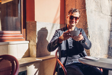 Portrait of stylish man relaxing in cafe outdoor drinking coffee. Guy looks at camera offering beverage on city street