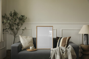 Grey textile sofa. Elegant living room interior. Blank vertical wooden picture frame poster mockup. Mediterranean, Scandinavian style. Cup of tea coffee, wool plaid. Table with marble lamp, olive tree