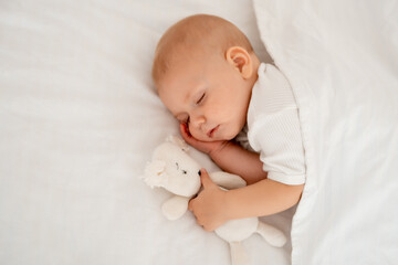 a newborn baby sleeps sweetly holding a toy in his hands, a place for text, a baby sleeps on a white bed under a blanket with a pen under his cheek, a healthy baby's sleep