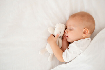 a newborn baby sleeps sweetly holding a toy in his hands, a place for text, a baby sleeps on a white bed under a blanket with a pen under his cheek, a healthy baby's sleep