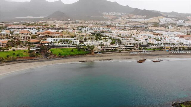 Aerial view of Costa Adeje city, Tenerife, Canary islands. Cinematic 4K footage with sandy coast, calm waters of Atlantic ocean, city modern villas, quay, mountains. Video during sandy storm calima