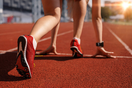 Female athlete at the starting line of a sprint race, wearing red track spikes, ready for competition on a sunlit outdoor stadium track.