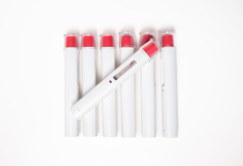 Top view of many pen injectors in a row with a red tip for biological treatments such as Adalimumab, Etanercept or epinephrine. The background is aseptic, clean, white and well lit.