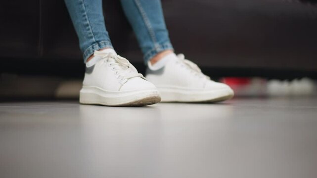 Close-up of lady's white canvas sneakers gently tapping left and right foot on floor with jean trousers while seated on a couch in a relaxed shopping mall setting