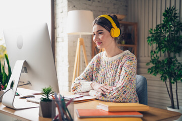 Young woman wearing yellow headphones studying at home while seated by a desktop computer in a cozy...