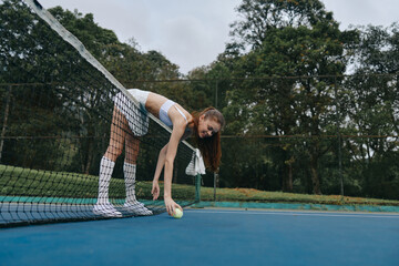 A young woman in sportswear picking up a tennis ball on a court surrounded by greenery, showcasing...