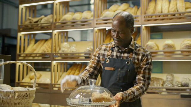 Friendly senior African American salesman in apron and gloves, handing paper bag of fresh croissants to female customer, then closing pastries on cake stand with lid at bakery counter