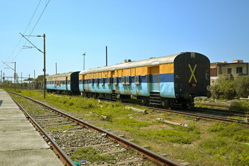 Fototapeta premium A stationary train carriage sits on a track in an open area. The carriage is painted blue and yellow, with barred windows and metal shutters.