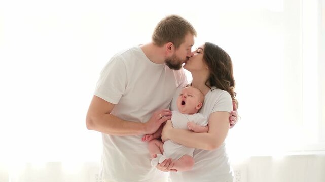 A happy young family mom dad and a small child smiling and kissing on a white isolated window background, a loving family threesome at home