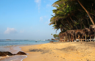 Tropical beach with golden sand, palm trees, and wooden huts by the sea, ideal for a summer vacation, tropical getaway, or relaxing holiday by the ocean