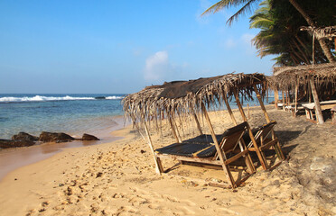 Tropical beach with wooden sun loungers under thatched umbrellas, surrounded by palm trees and golden sand, perfect for a relaxing summer holiday by the sea