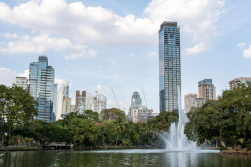 A tranquil pond features a graceful fountain amidst lush greenery, with modern skyscrapers rising in the background under a beautiful sky, inviting calm and serenity in bustling Bangkok