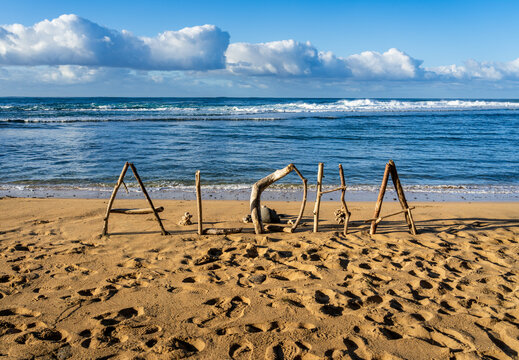 The word 'Aloha' is spelled out using driftwood and other natural materials on a sandy Kauai beach with the ocean in the background.