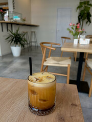 A glass of iced coffee with a frothy top and a black straw sits on a wooden table in a modern cafe with wooden chairs, plants, and minimal decor