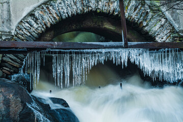 Frozen icicles hang over rushing water beneath a stone arch in Sweden during winter