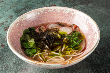 A delicious bowl of ramen noodles topped with fresh herbs, seaweed, radishes, and green onions, served in a beautifully textured ceramic bowl on a dark background.
